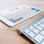 Palabras más buscadas en los buscadores A modern tablet displaying a search engine logo next to a wireless keyboard on a wooden desk.