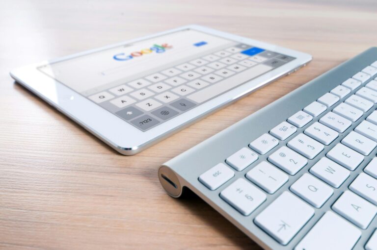 Palabras más buscadas en los buscadores A modern tablet displaying a search engine logo next to a wireless keyboard on a wooden desk.